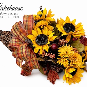 May include: A woven cornucopia filled with artificial yellow sunflowers, dark red berries, green leaves, and a pinecone. A plaid ribbon in shades of orange, yellow, and brown is tied to the side. The text Lakehouse BOWTIQUE is visible at the top.