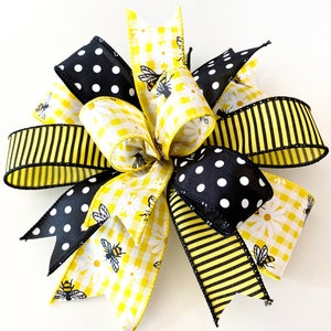 May include: A yellow and black bow with a gingham pattern, black polka dots, and a bee and daisy print. The bow is tied with a black ribbon.