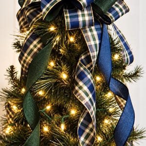 May include: A large decorative bow topper for a Christmas tree featuring layered ribbons in navy blue, forest green, and a blue-green plaid pattern. The bow has long, cascading ribbon tails draped down the lit evergreen branches.