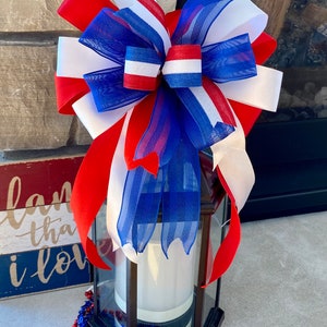 May include: A red, white and blue bow made of ribbon and mesh is tied around a black lantern. The lantern is sitting on a concrete surface with red, white and blue beads scattered around it.