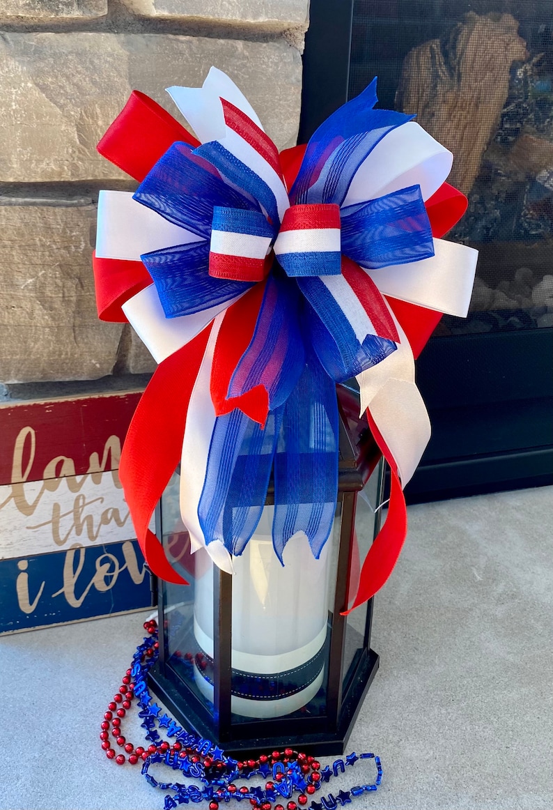 May include: A red, white and blue bow made of ribbon and mesh is tied around a black lantern. The lantern is sitting on a concrete surface with red, white and blue beads scattered around it.