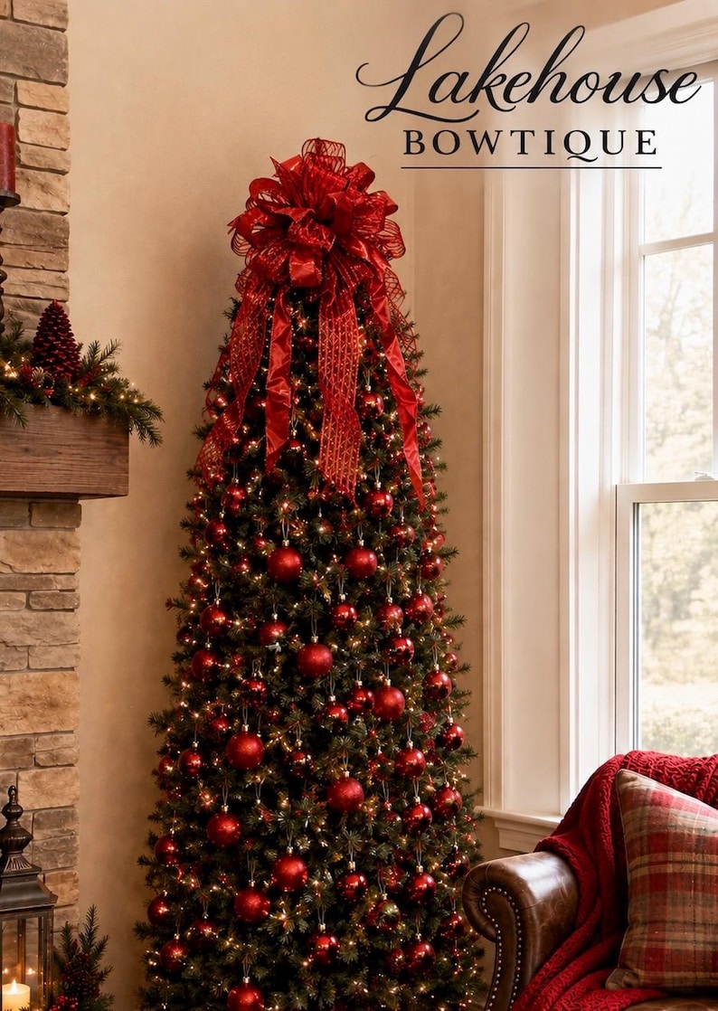May include: A tall, slender Christmas tree decorated with numerous shiny red spherical ornaments and warm white string lights. A large, multi-layered red ribbon bow with a textured mesh pattern sits at the very top. Text reads Lakehouse BOWTIQUE.