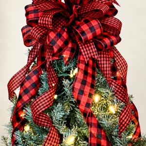 May include: A large decorative bow made of red and black buffalo plaid ribbon, featuring multiple loops and long trailing tails. The bow is positioned atop a green artificial Christmas tree illuminated with warm white string lights. Text reads Lakehouse BOWTIQUE.