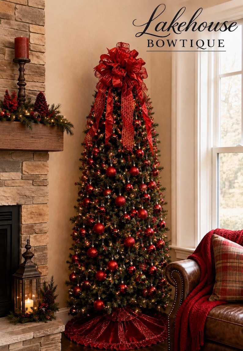 May include: A tall, slim Christmas tree decorated with numerous shiny red spherical ornaments and warm white string lights. A large, elaborate red ribbon bow sits at the top, and a ruffled red fabric tree skirt covers the base. Text reads Lakehouse BOWTIQUE.