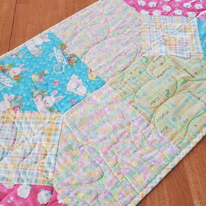 以下が含まれることがあります： ウサギ、子羊、花柄のパッチワークデザインが施されたキルトのテーブルランナー。ランナーはコットン生地で作られており、薄い青、ピンク、緑の色合いです。生地には「春」、「喜び」、「色」、「幸せ」などの文字が印刷されています。