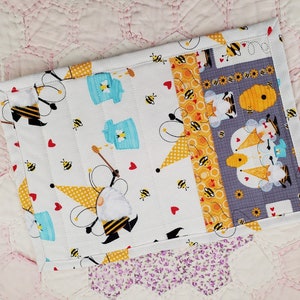 May include: A white fabric pot holder with a bee and honey pot theme. The pot holder is made of quilted fabric and has a gray border. The fabric features bees, honey pots, hearts, and gnomes.