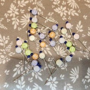 以下が含まれることがあります： ガラス製の頭部を持つカラフルな裁縫針のセット。頭部は青、緑、オレンジ、紫の色合いです。針は花柄の布地に並べてあります。