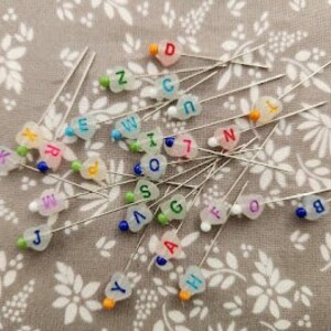 May include: Assortment of colorful alphabet letter pins with silver metal shafts. The letters include A, B, D, E, G, H, I, J, K, L, M, N, O, R, S, U, V, W, X, Y, and Z.