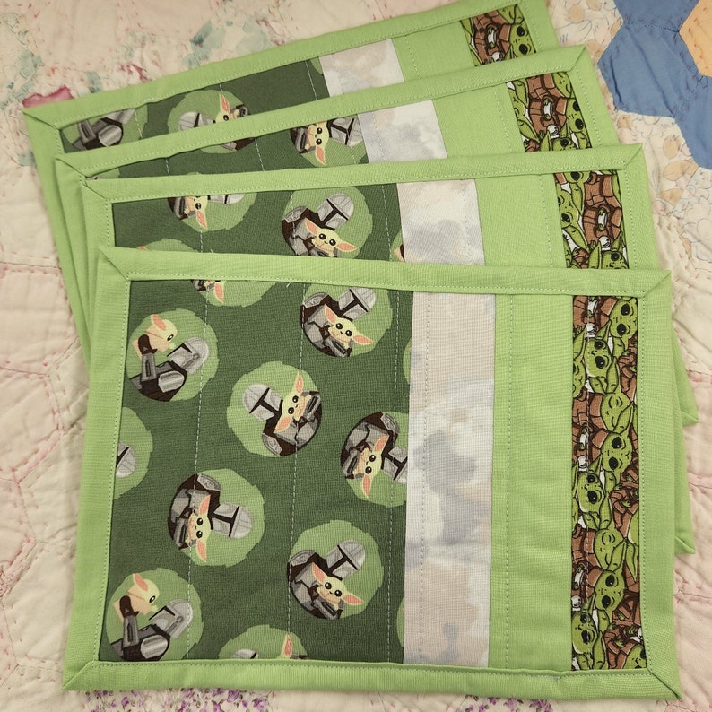 Puede incluir: Juego de cuatro manteles individuales de tela verde con borde blanco y un patr&oacute;n repetido de un personaje de dibujos animados. El personaje es verde y tiene ojos y orejas grandes.