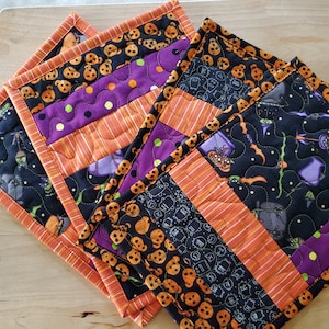 May include: Three quilted fabric pot holders with a Halloween theme. The pot holders feature a black background with orange, purple and white accents. One pot holder has a pattern of pumpkins, another has a pattern of bats and ghosts, and the third has a pattern of spiders and webs.