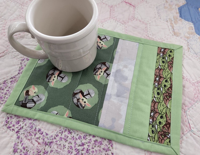 Puede incluir: Posavasos de tela verde y blanco con un patr&oacute;n repetido de un personaje de dibujos animados con casco. El posavasos tiene un borde blanco y verde y est&aacute; sobre una colcha rosa y blanca.