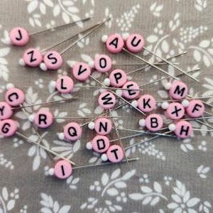 以下が含まれることがあります： ピンクと白のアルファベットの文字が書かれた縫い針のセットです。文字は黒で、針の上部に書かれています。針はランダムな順序で配置されています。