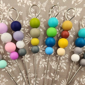 Puede incluir: Un conjunto de diez agitadores de metal y cuentas de silicona de colores vivos. Las cuentas están dispuestas en un patrón de arcoíris con una variedad de colores, incluyendo rosa, azul, verde, amarillo, rojo, morado y gris. Los agitadores de metal son plateados y tienen un bucle en la parte superior.