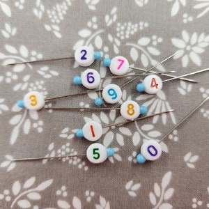 May include: Ten white plastic sewing pins with colorful numbers on the heads. The pins are arranged on a gray fabric with a white floral pattern.