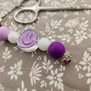 May include: A purple, white, and blue beaded keychain with a clear charm featuring a smiley face. The keychain also has a small gold heart charm.