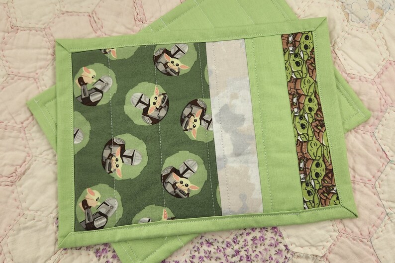 Puede incluir: Dos posavasos de tela verde con un dise&ntilde;o acolchado. Un lado presenta un patr&oacute;n repetido de un peque&ntilde;o personaje verde con casco. El otro lado tiene un borde verde con un patr&oacute;n repetido del mismo personaje.