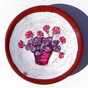 Può includere: Piatto decorativo rotondo dipinto a mano, con bordo rosso e interno bianco. Il piatto presenta un vaso rosa con fiori viola e rosa. L'opera è firmata con le iniziali "GM" nell'angolo in basso a sinistra.