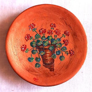 Può includere: Piatto in terracotta dipinto a mano con un motivo floreale. Il piatto è di colore arancione caldo, con un'immagine centrale di un vaso marrone pieno di foglie verdi e fiori rossi. Il piatto ha un aspetto rustico e fatto a mano.