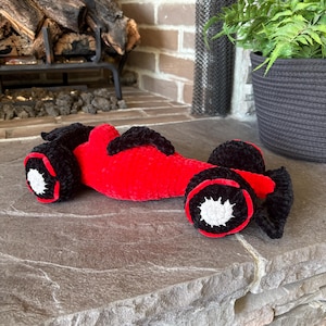 May include: A handmade, red and black crochet race car toy. The toy features black wheels with white and red accents. The body of the car is a vibrant red, and the overall design is soft and plush.