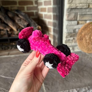 May include: A crocheted toy race car made of bright pink fuzzy yarn. The vehicle features four black wheels with white centers, a raised cockpit area, and a rear spoiler. The soft, textured construction mimics the shape of a formula-style racing car.