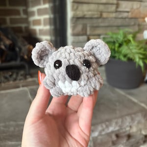May include: A gray, plush koala toy with black eyes and a black nose. The koala has small, rounded ears and a white patch under its nose. The toy is made of a soft, textured material.