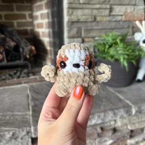 May include: A handmade, plush sloth toy in shades of tan, brown, and white. The sloth has small arms and a round body. The eyes are black, and the toy is made of a soft, textured material. The sloth is a cute, cuddly toy.