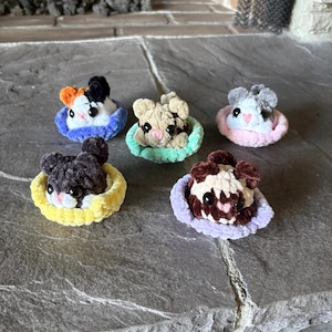 May include: Five small, crocheted plush animals, each sitting in a soft, circular base. The animals feature fuzzy yarn textures, black bead eyes, and small pink noses. They are colored in various patterns of white, black, brown, and orange.