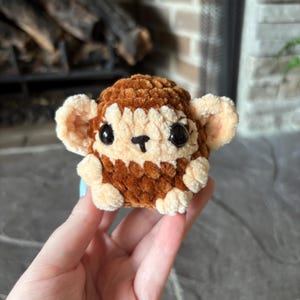 May include: A handmade, plush monkey toy in shades of brown and cream. The monkey has large, black eyes, a small black nose, and cream-colored ears. The toy is made of soft, textured yarn.