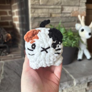 May include: A handmade crochet cat toy in white, orange, and black. The cat has black button eyes, a black stitched mouth, and a black 'X' for a nose. The cat has a small black tail and is held in a hand.