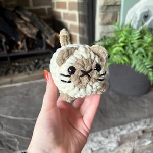 May include: A small, crocheted cat figurine in cream and tan. The cat has black eyes, whiskers, and a stitched mouth. The handmade plush toy is held in a hand, with a fireplace and greenery in the background.