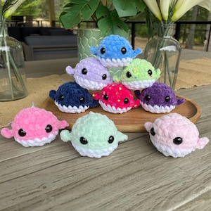 May include: A collection of nine crocheted plush pufferfish toys in various colors, including pink, blue, green, purple, and red. The toys have white crocheted mouths and black eyes.