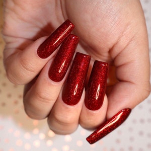 May include: Close-up of a hand with five long, red glitter nails. The nails are all the same length and shape, and they are painted with a bright red glitter polish.