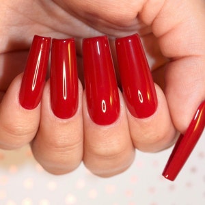 May include: A hand showing five long, rectangular, glossy red nails. The nails are a vibrant, solid colour, with a smooth, reflective surface. One nail is detached and resting beside the others. The background is a soft, blurred pattern.