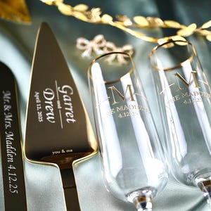 May include: Gold wedding cake server and two champagne flutes with personalized engraving. The server has the text "Garret and Drew" and "April 12, 2025". The flutes have the text "M" and "The Maddens 4.12.25".