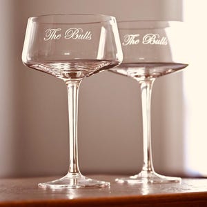 May include: Two clear glass coupe glasses with long, slender stems and wide, shallow bowls. Each glass features the engraved text The Bulls in a cursive script font on the side of the bowl, showcasing a transparent design and smooth finish.