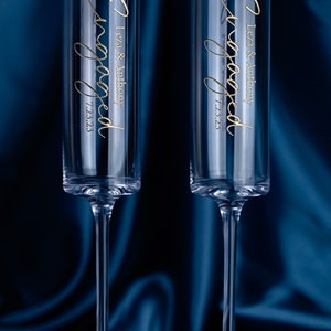 May include: Two clear glass champagne flutes with gold lettering that reads "Engaged" with the names "Leza & Anthony" and the date "7.23.23".
