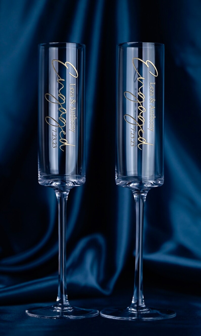 May include: Two clear glass champagne flutes with gold lettering that reads "Engaged" with the names "Leza & Anthony" and the date "7.23.23".