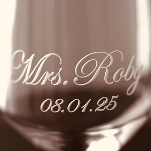 Puede incluir: Primer plano de una copa de champ&aacute;n con la inscripci&oacute;n "Mrs. Roby" y la fecha "08.01.25" grabadas en una fuente elegante. El cristal tiene un tono sepia, lo que sugiere un tema vintage o rom&aacute;ntico.