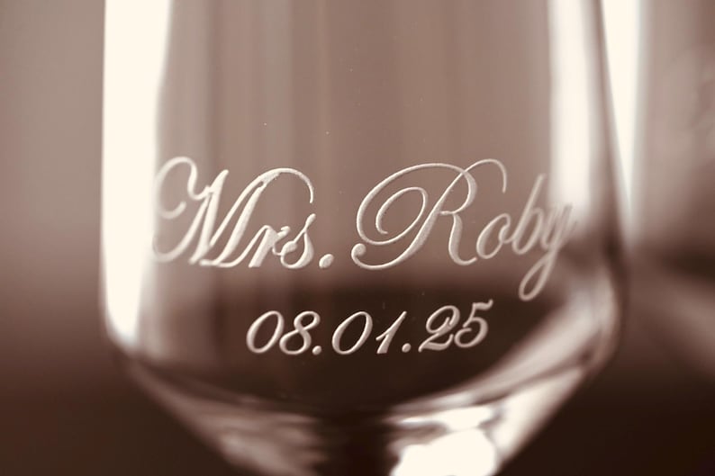 Puede incluir: Primer plano de una copa de champ&aacute;n con la inscripci&oacute;n "Mrs. Roby" y la fecha "08.01.25" grabadas en una fuente elegante. El cristal tiene un tono sepia, lo que sugiere un tema vintage o rom&aacute;ntico.