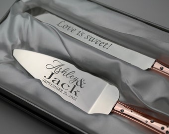 May include: A silver cake knife and server set with rose gold handles. The knife has a serrated edge and the server has a rounded edge. The knife is engraved with "Love is sweet!" and the server is engraved with "Ashley & Jack SEPTEMBER 25, 2022".