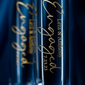 May include: Two clear glass champagne flutes with gold lettering that reads "Engaged" and "Leza & Anthony 7.23.23".