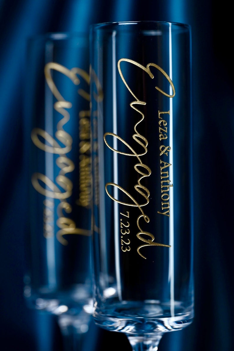 May include: Two clear glass champagne flutes with gold lettering that reads "Engaged" and "Leza & Anthony 7.23.23".