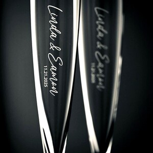 May include: Two clear glass champagne flutes featuring vertical etched text that reads Linda & Eamon followed by the date 11.21.2025. The glasses have a tall, slender, tapered silhouette with a smooth, reflective surface against a dark background.
