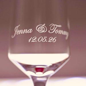 Puede incluir: Una copa de champ&aacute;n transparente con los nombres "Jenna & Tommy" y la fecha "12.05.26" grabados en una elegante escritura. La copa tiene un tallo largo y una copa ancha, ideal para una boda o aniversario. Medidas en cent&iacute;metros.