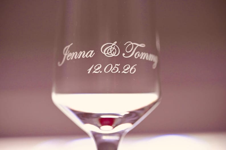 Puede incluir: Una copa de champ&aacute;n transparente con los nombres "Jenna & Tommy" y la fecha "12.05.26" grabados en una elegante escritura. La copa tiene un tallo largo y una copa ancha, ideal para una boda o aniversario. Medidas en cent&iacute;metros.