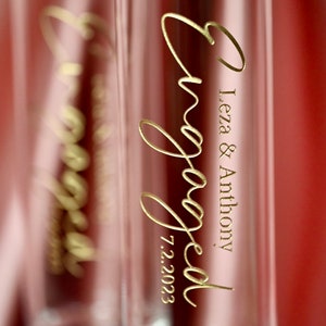 May include: Two clear glass champagne flutes with gold lettering that reads "engaged" and "Leza & Anthony 7.2.2023".