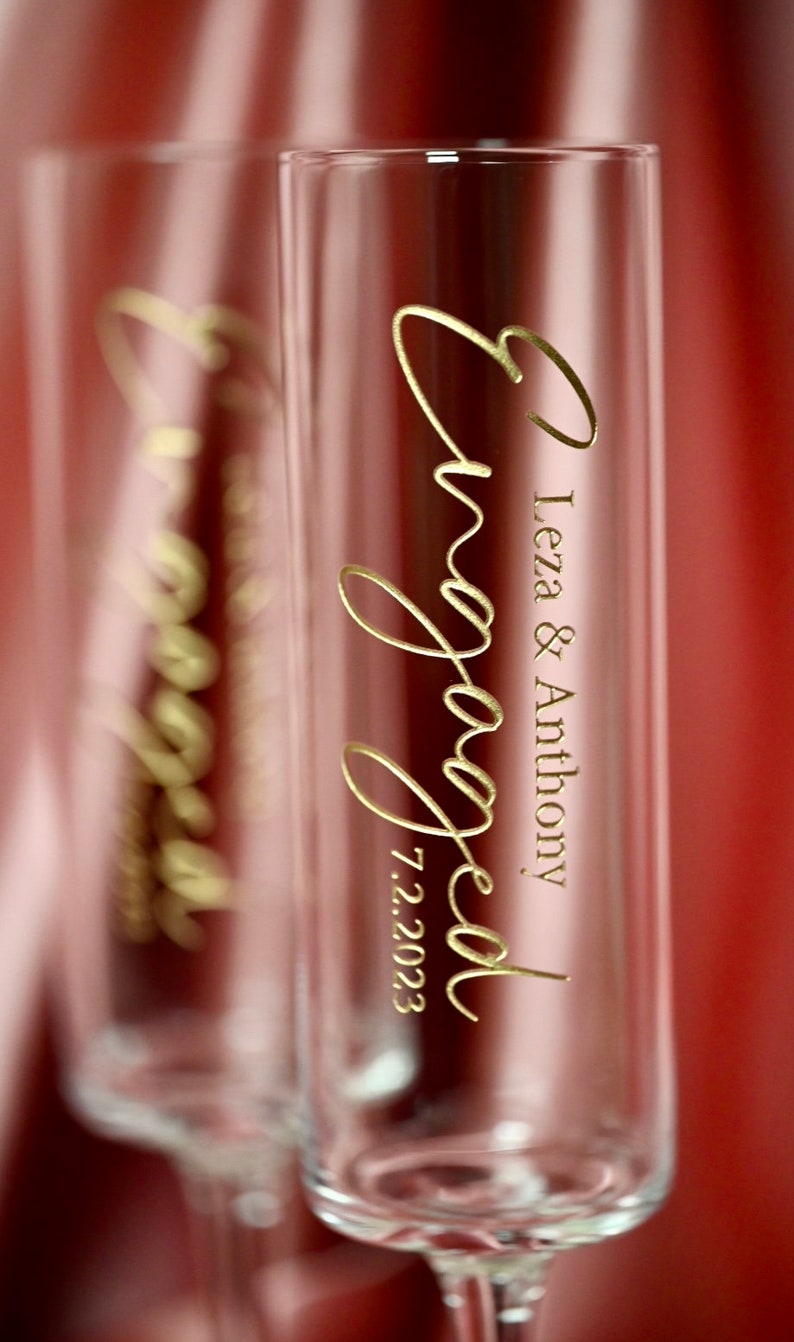 May include: Two clear glass champagne flutes with gold lettering that reads "engaged" and "Leza & Anthony 7.2.2023".