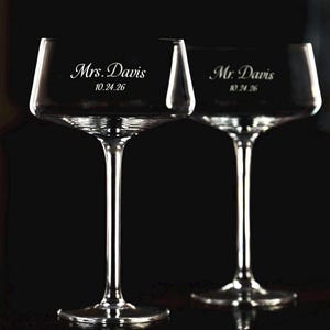 May include: Two clear glass coupe glasses with long stems and wide bowls. One glass is etched with the text Mrs. Davis and the date 10.24.26. The second glass is etched with the text Mr. Davis and the date 10.24.26. The glasses are transparent.