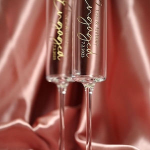 May include: Two clear champagne flutes with gold lettering that says "Engaged" and the date "7.2.2023". One flute says "Leza & Anthony" and the other says "Tess & William". The flutes are on a pink satin background.