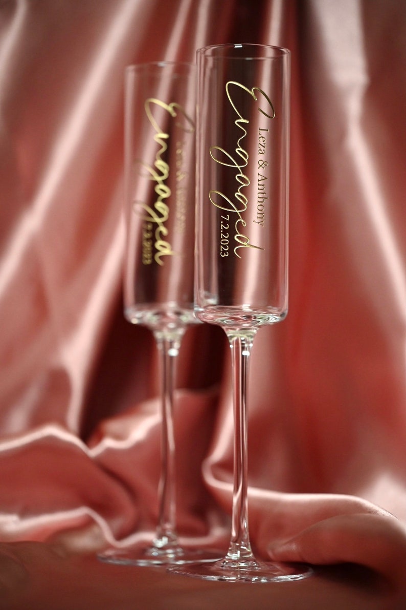 May include: Two clear champagne flutes with gold lettering that says "Engaged" and the date "7.2.2023". One flute says "Leza & Anthony" and the other says "Tess & William". The flutes are on a pink satin background.
